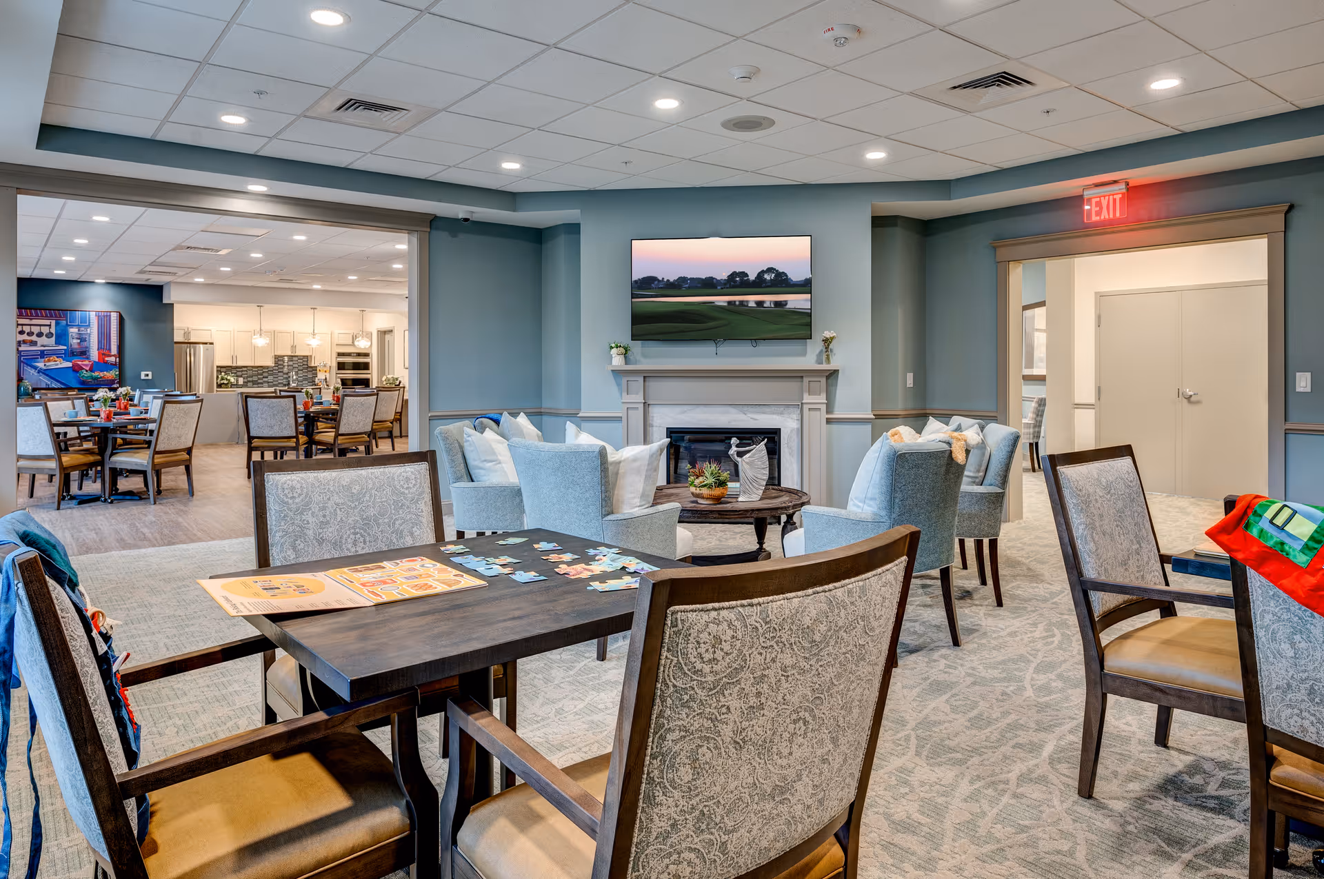 A cozy common area in a senior living facility featuring a seating arrangement with four light blue armchairs around a round wooden coffee table in front of a fireplace with a mounted flat-screen TV above it. In the foreground, there is a dark wooden table with four upholstered chairs, some puzzle pieces, and a booklet on the table. The background shows an open dining area with tables and chairs, a kitchen with white cabinetry, and modern lighting fixtures. The walls are painted in soft blue tones, and the ceiling has recessed lighting. An exit sign is visible above a doorway on the right.