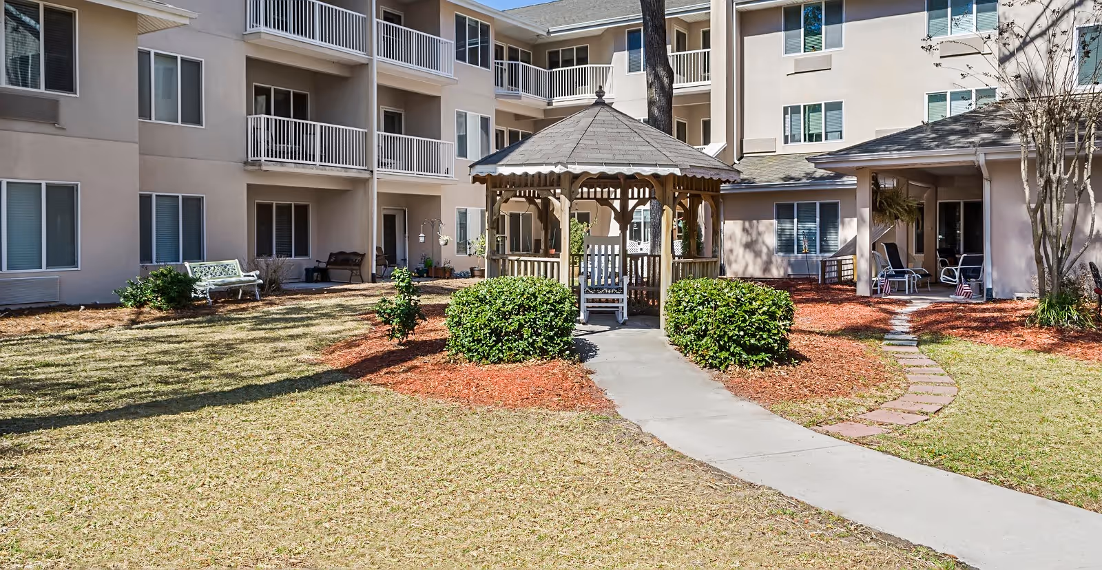Outdoor courtyard area of a senior living facility with a wooden gazebo surrounded by bushes and a concrete pathway leading to it. The building has multiple balconies and windows, with benches and chairs placed around the courtyard.