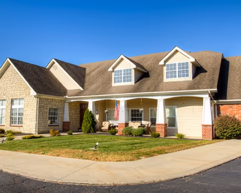 Residential-style senior living building with a covered front porch, dormer windows, and a manicured lawn.