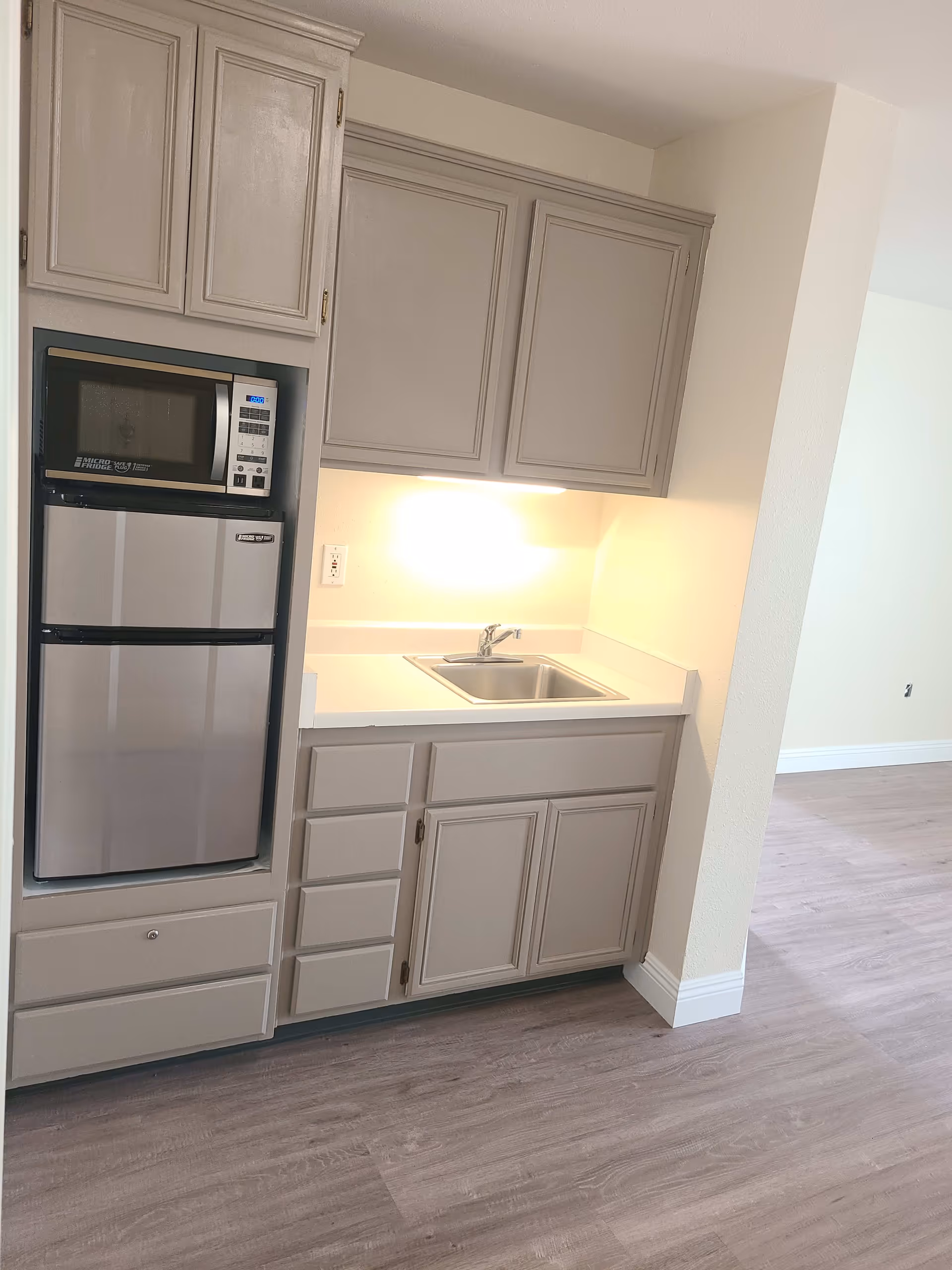 Small kitchenette area with light gray cabinets, a stainless steel microwave and mini refrigerator, a single basin sink with a faucet, and under-cabinet lighting. The floor is light wood laminate and the walls are painted a light cream color.