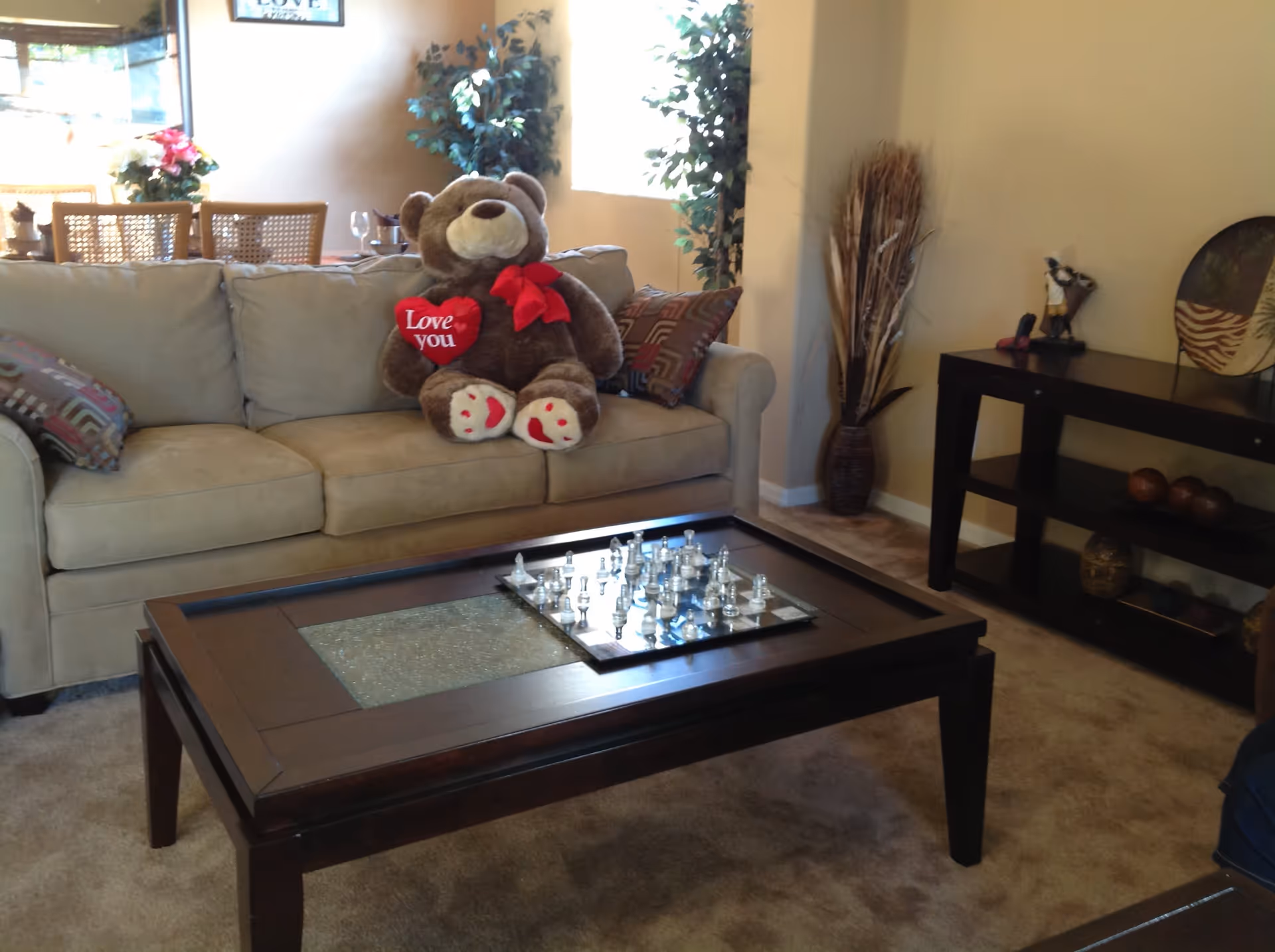 A cozy living room with a beige sofa adorned with patterned pillows and a large teddy bear holding a red heart that says 'Love you'. In front of the sofa is a dark wooden coffee table with a glass inset and a chess set on top. The room has beige walls, a carpeted floor, a decorative plant in the corner, and a dark wooden console table with decorative items on the right side.