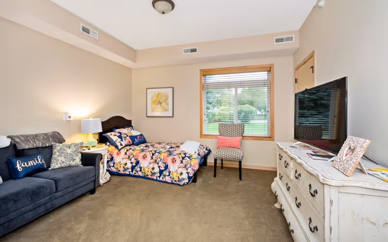 A cozy bedroom in an assisted living facility featuring a floral bedspread on a single bed, a dark blue couch with decorative pillows including one that says 'family', a white nightstand with a lamp, a patterned chair with a pink cushion near a window showing greenery outside, and a white dresser with a flat-screen TV and picture frames on top.