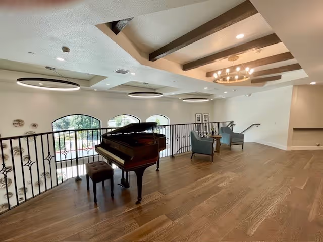 Spacious interior area with wooden flooring featuring a grand piano and a piano bench near a black metal railing. Two blue armchairs and a small wooden table are placed near the wall under a ceiling with exposed wooden beams and modern circular light fixtures. Large arched windows provide natural light and a view of greenery outside.