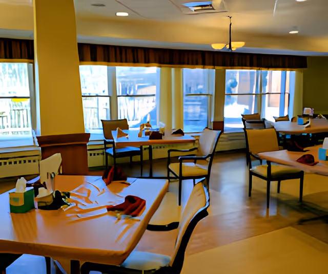A dining room with several tables and chairs arranged neatly. Each table has napkins and tissue boxes placed on top. Large windows with brown valances allow natural light to fill the room.