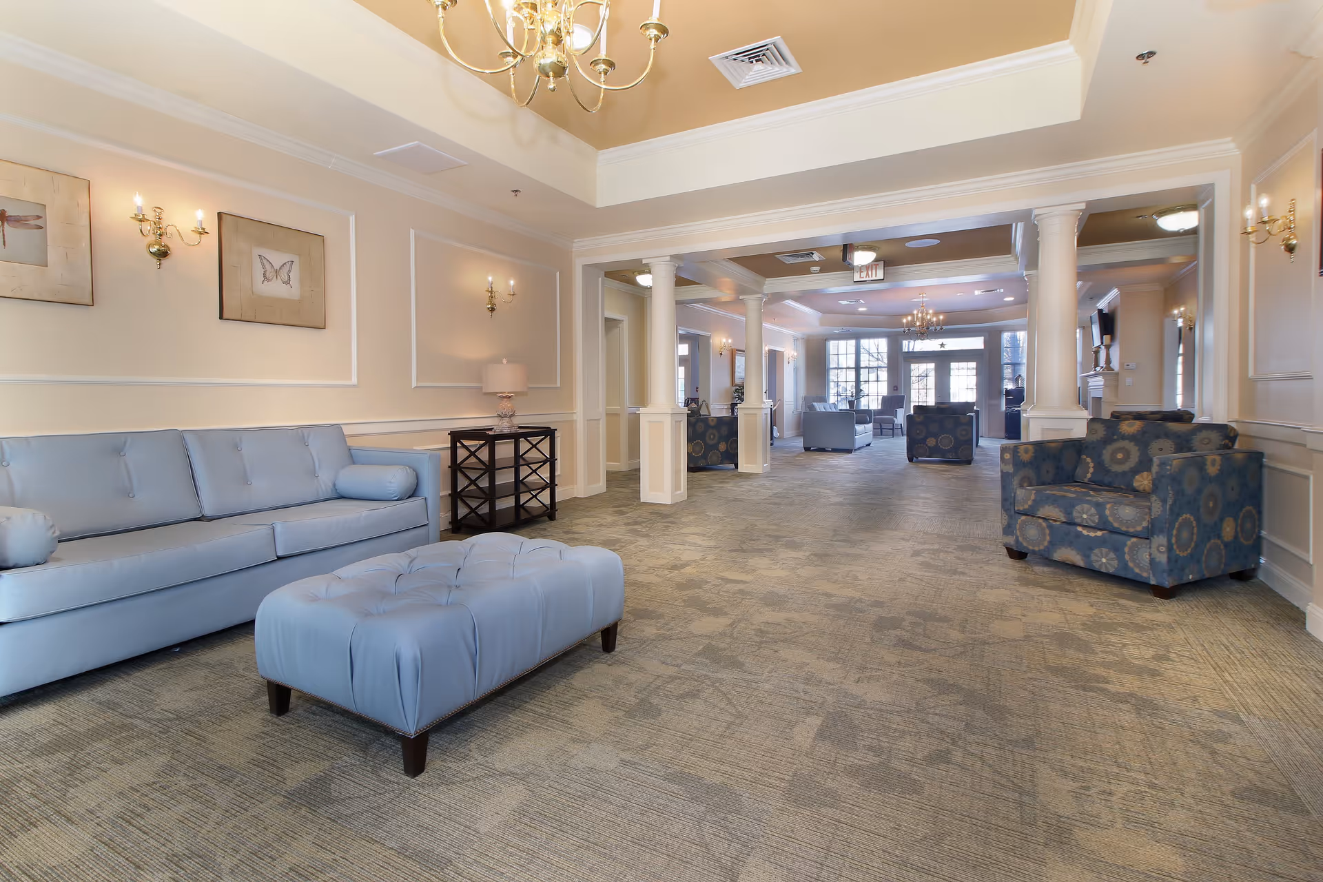 Spacious and well-lit senior living facility lounge area with light blue sofas, a matching ottoman, patterned armchairs, beige walls with framed butterfly artwork, wall sconces, and chandeliers. The room features carpeted flooring, white columns, and large windows letting in natural light.
