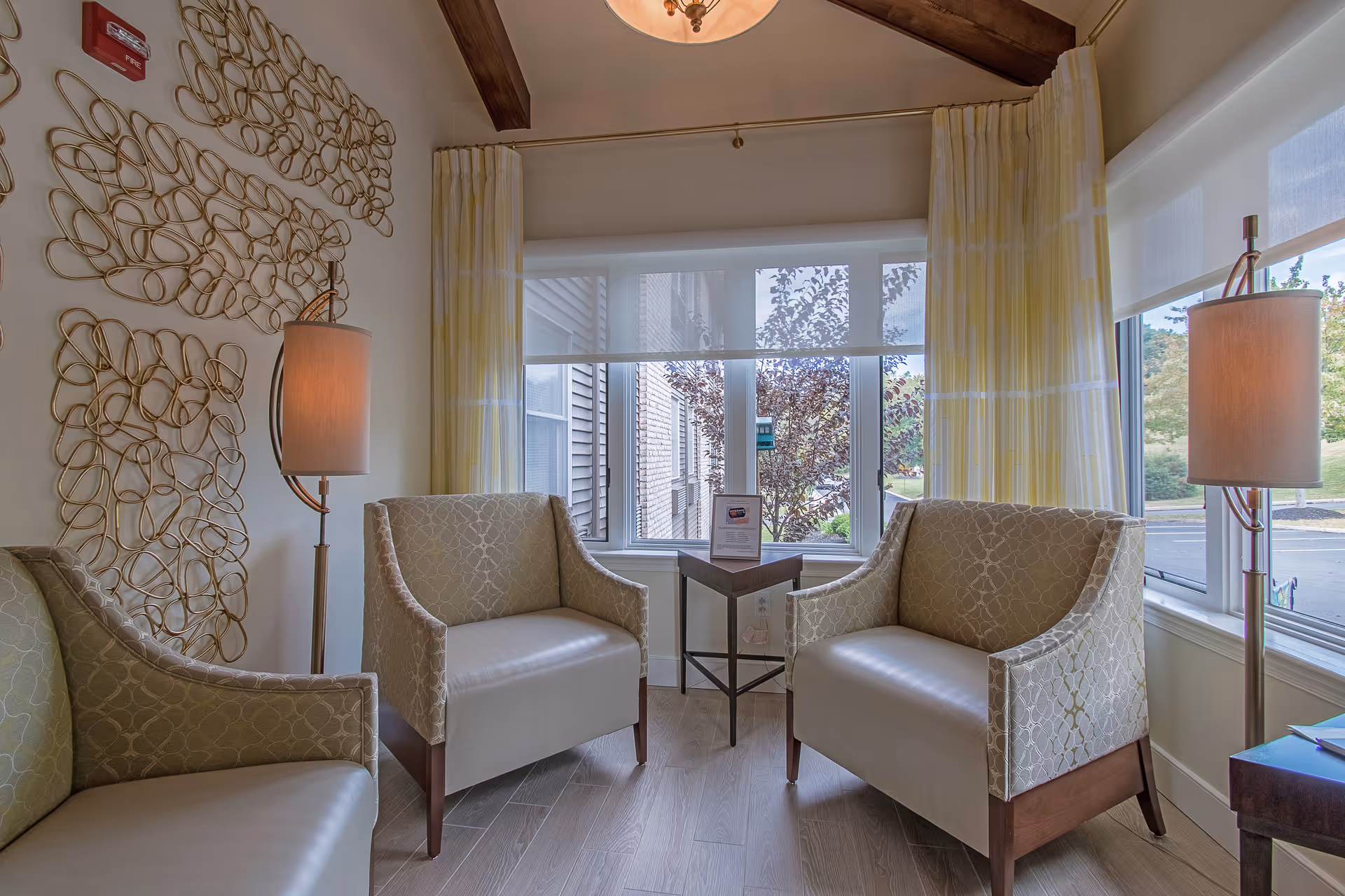 A bright sitting area with two upholstered armchairs, a small side table and floor lamps in front of a bay window with curtains and decorative wall art.