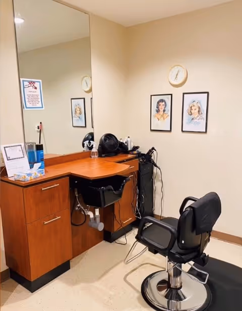 A small hair salon station with a black salon chair, a wooden counter with drawers, a large mirror, a black sink, and hair styling tools. Two framed portraits and a clock are mounted on the beige wall.