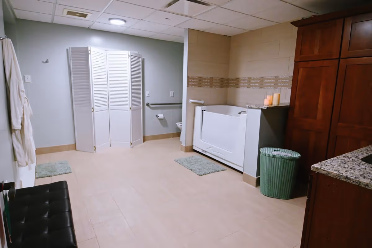 A spacious bathroom with beige tiled floor and walls, featuring a walk-in bathtub with grab bars, a toilet partially visible behind a white folding privacy screen, a green laundry basket, wooden cabinets with granite countertop, two bath mats, and a white bathrobe hanging on the wall.