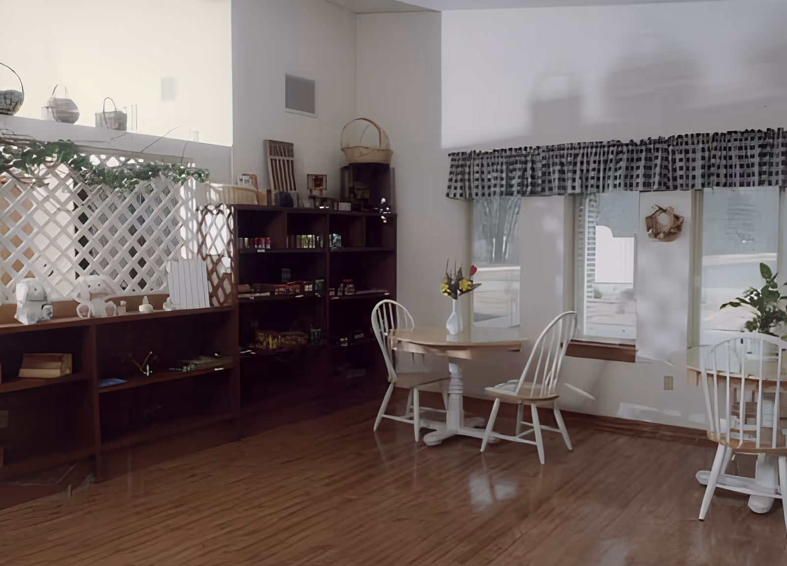 A cozy interior space with wooden flooring featuring a small round table with two white wooden chairs. A vase with flowers is placed on the table. To the left, there is a dark wooden shelving unit with various items and a white lattice divider with hanging baskets on top. The windows have checkered valances, and natural light is coming through.