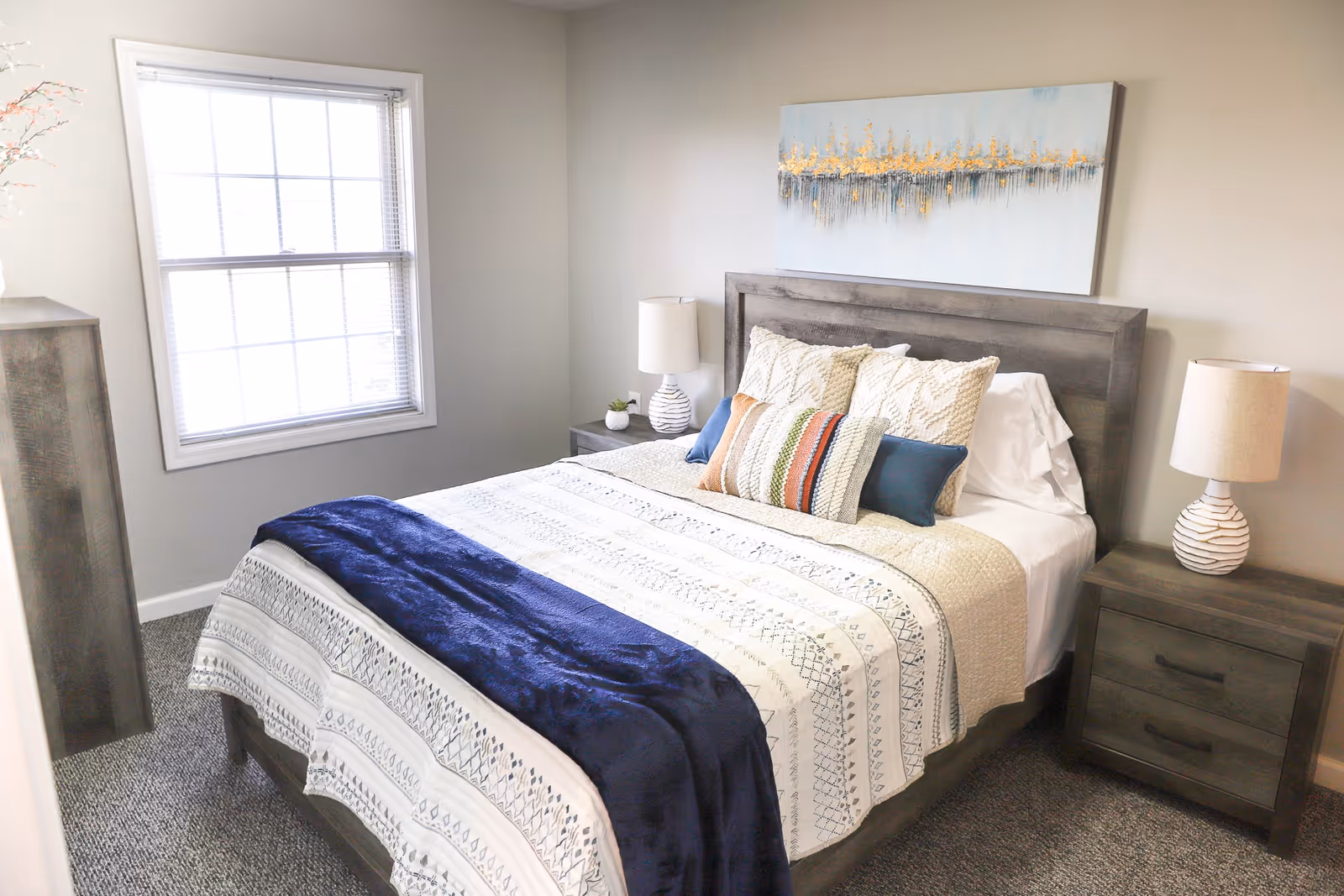 A neatly made bed with a wooden headboard in a bright bedroom. The bed has multiple pillows in various colors and patterns, a white and patterned bedspread, and a dark blue throw blanket. There are two matching wooden nightstands on either side of the bed, each with a lamp. A large window with blinds allows natural light into the room, and a decorative painting hangs above the headboard.