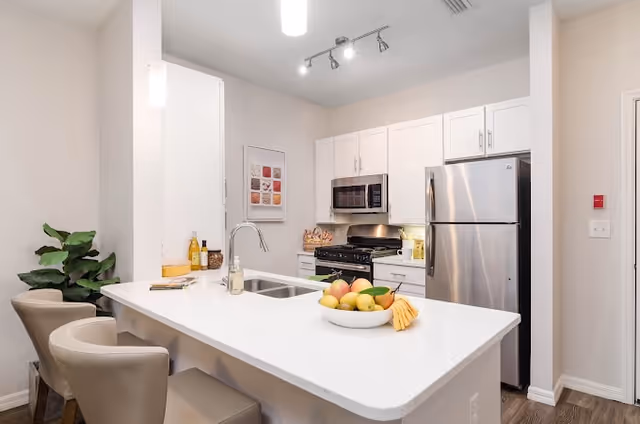 Modern kitchen with white cabinets, stainless steel refrigerator and microwave, a gas stove, and a white countertop island with a sink. The island has three beige chairs and a bowl of assorted fruits. There is a green plant in the corner and a framed picture on the wall.