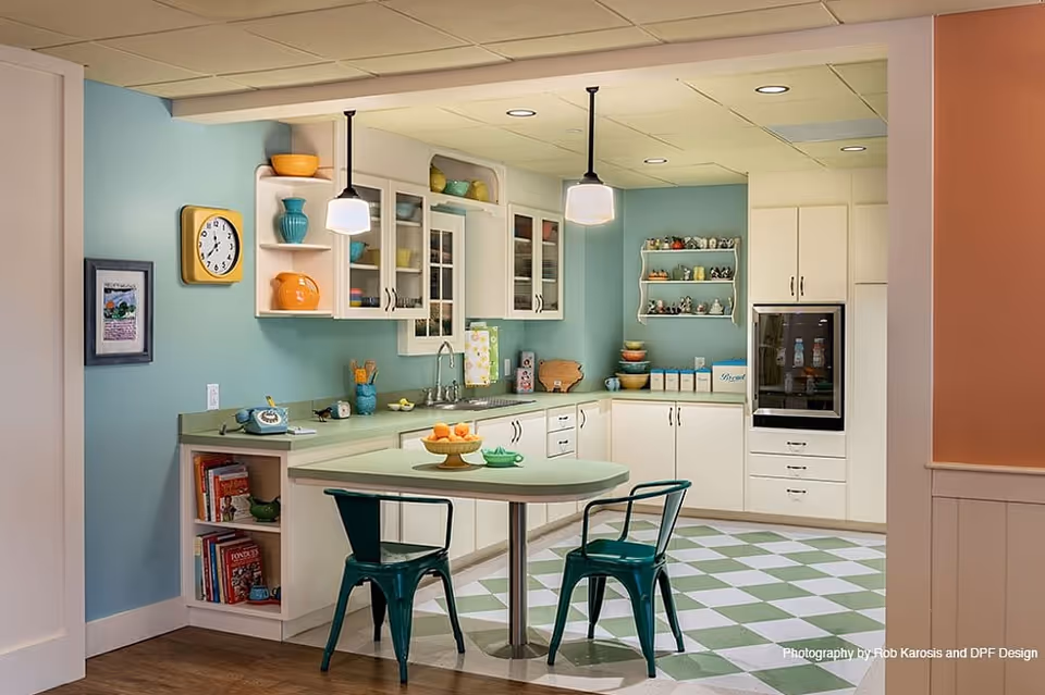 A retro-style kitchen with light blue walls, white cabinets, and a green and white checkered floor. The kitchen features a small table with two green metal chairs, pendant lights hanging from the ceiling, a wall clock, open shelves with colorful dishes, and a built-in oven.