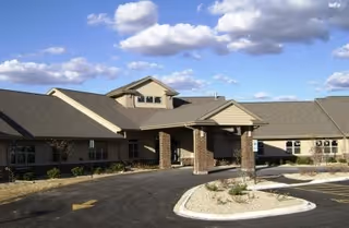 Single-story brick and siding senior living facility with a covered entrance, circular driveway, and landscaped islands under a partly cloudy sky.