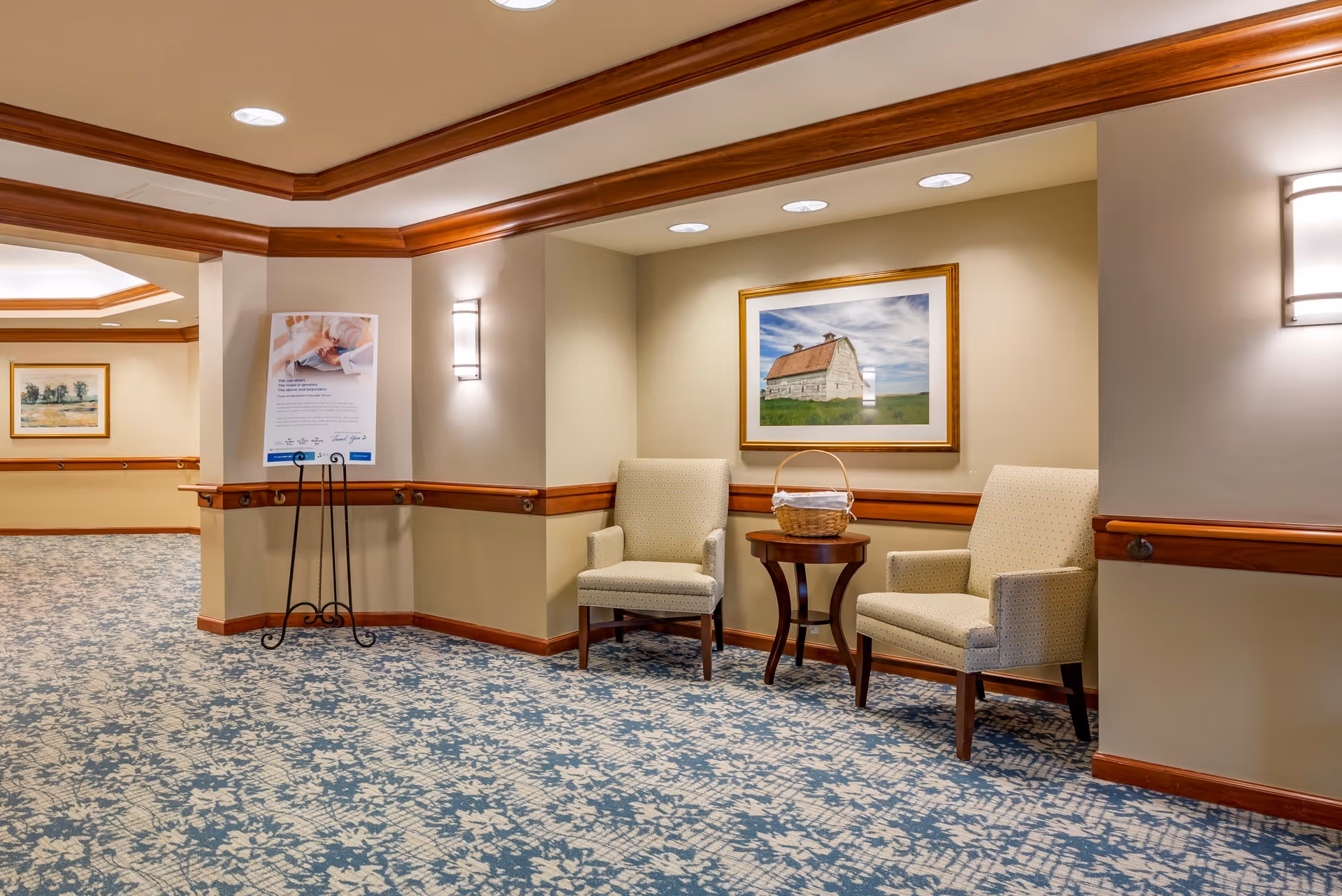A hallway corner in a senior living facility with two beige upholstered armchairs and a small wooden table between them holding a wicker basket. The walls are painted light beige with wooden trim and handrails. A framed picture of a barn hangs on the wall above the chairs. The carpet has a blue and white floral pattern. There is a poster on a black metal stand in the hallway and wall-mounted light fixtures provide illumination.