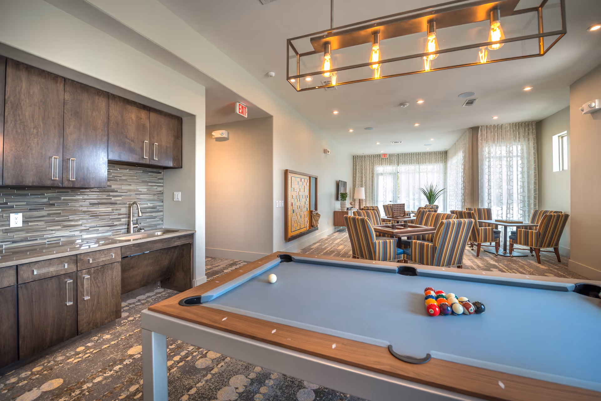 A recreational room with a pool table in the foreground, a kitchenette with dark wood cabinets and a sink on the left, and a seating area with striped armchairs and tables near large windows with sheer curtains in the background. The room is well-lit with ceiling lights and a modern rectangular chandelier above the pool table.