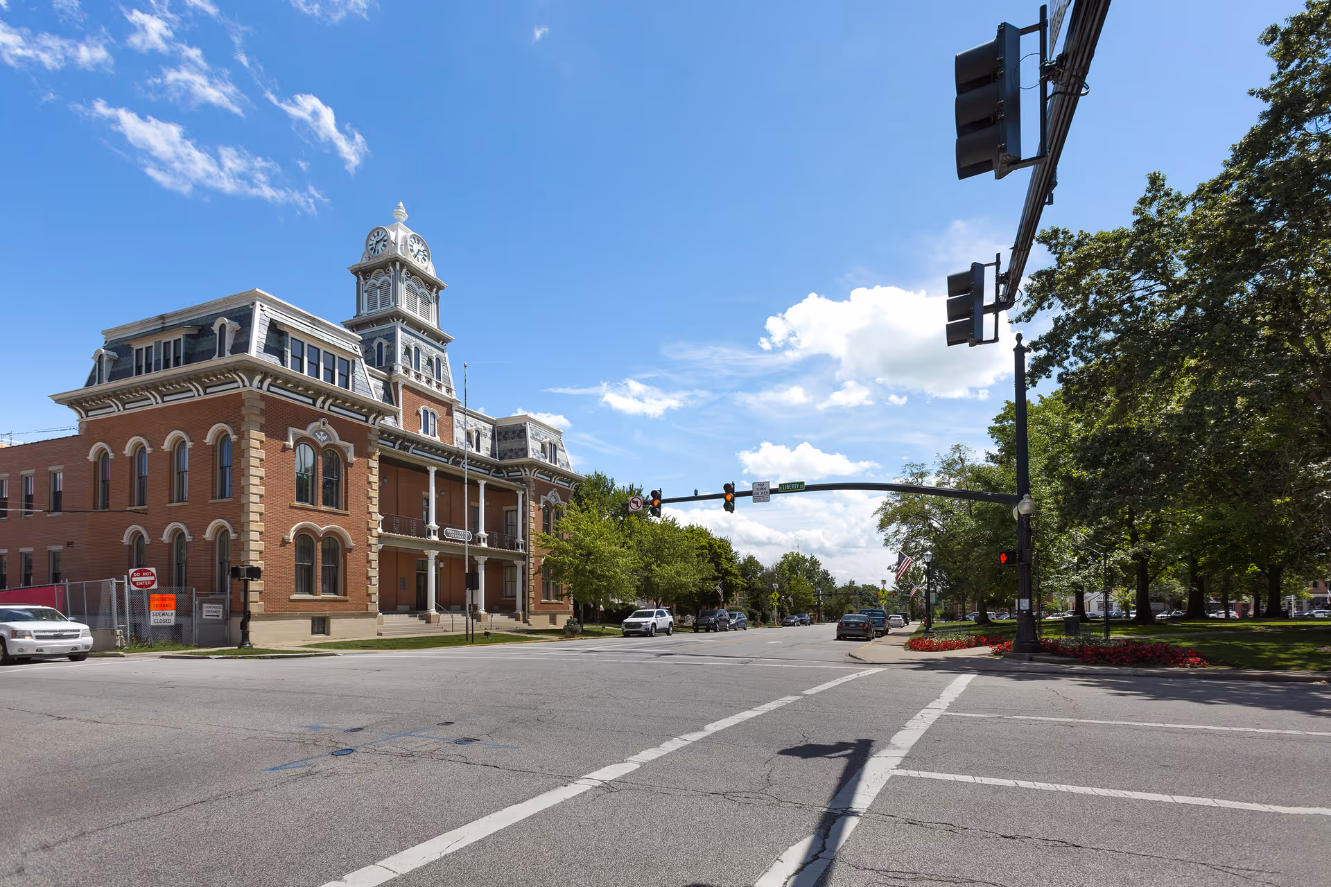 Historic three-story brick building with a clock tower at a tree-lined street intersection under a blue sky.