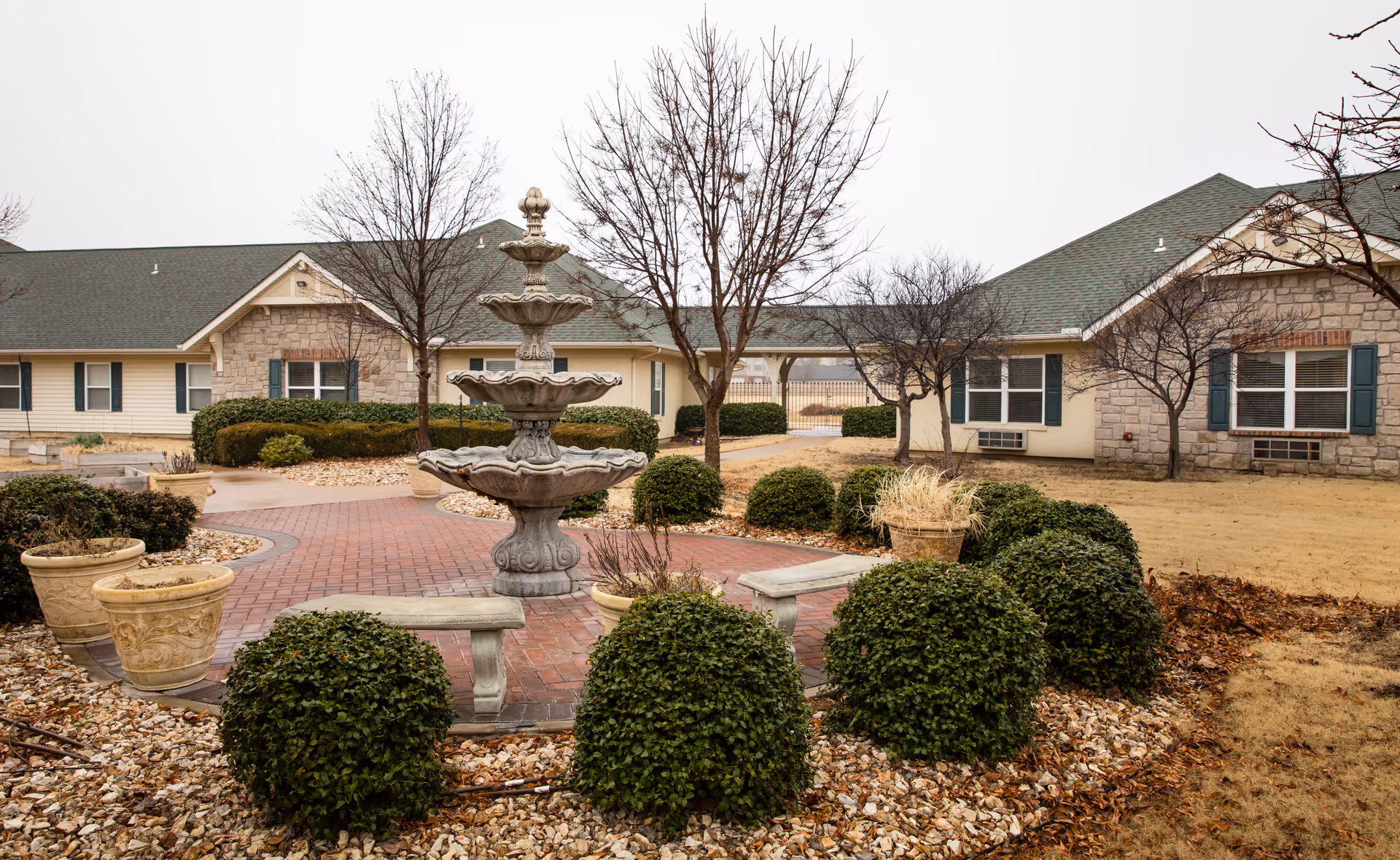 Outdoor courtyard area with a multi-tiered stone fountain in the center, surrounded by neatly trimmed bushes and stone benches. The courtyard is paved with red bricks and has several large decorative planters. In the background, there are single-story buildings with green roofs, beige siding, and stone accents. Leafless trees are scattered throughout the area, indicating a winter or early spring season.