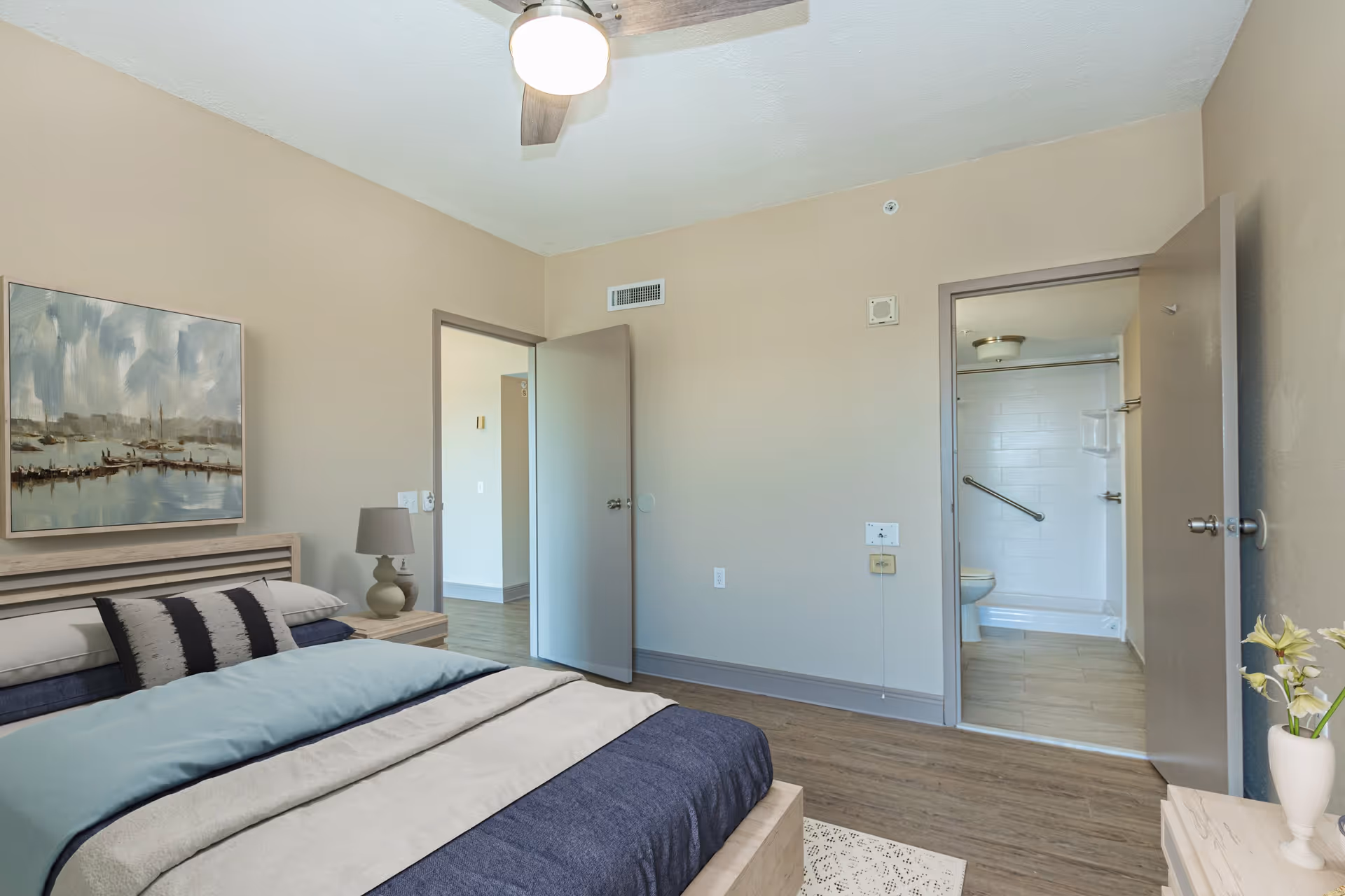 A bedroom with a neatly made bed featuring blue and beige bedding, a nightstand with a lamp, a painting of a harbor scene on the wall, and two open doors showing a hallway and a bathroom with a shower and grab bar.