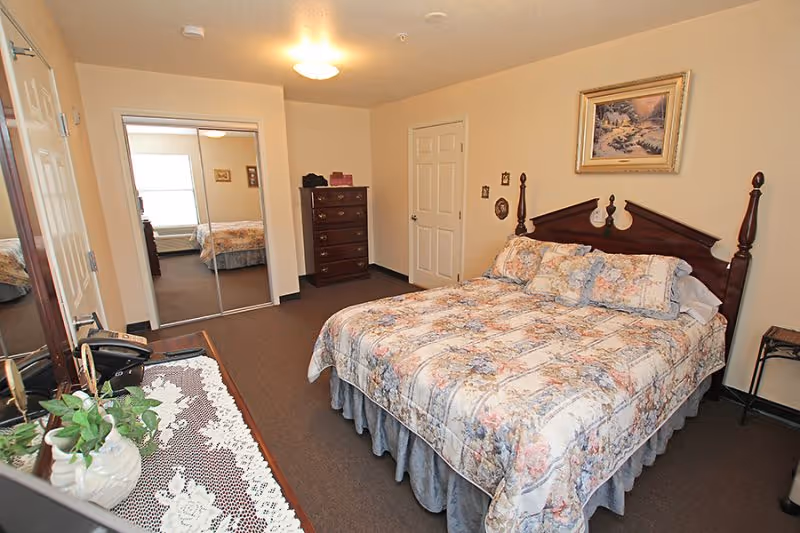 A cozy bedroom in a senior living facility featuring a large bed with floral bedding, a wooden headboard, a dresser with drawers, a closet with mirrored sliding doors, a small side table, and a framed painting on the wall. The room has beige walls and carpeted flooring.