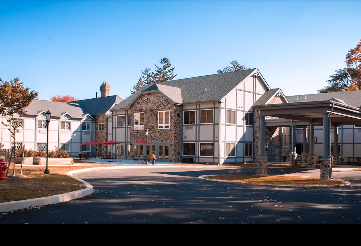 Exterior view of a senior living facility building with stone and white panel walls, a covered entrance, outdoor seating with red umbrellas, and a curved driveway in front.