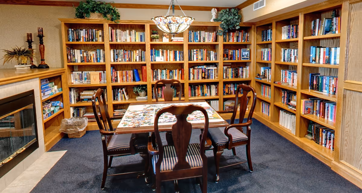 A cozy library room with wooden bookshelves filled with books lining the walls. In the center, there is a wooden table with a partially completed jigsaw puzzle on it, surrounded by four wooden chairs with striped cushions. A decorative hanging lamp is above the table, and a fireplace is visible on the left side of the room.