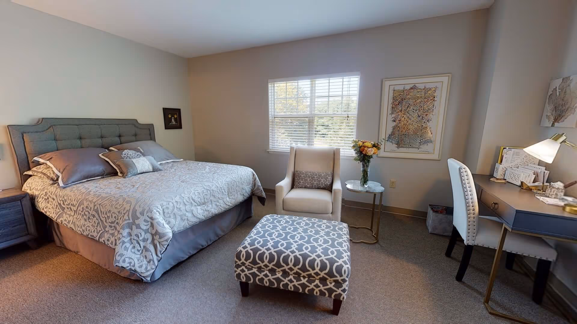 A cozy bedroom with a large bed featuring a tufted headboard and patterned bedding. There is a beige armchair with a decorative pillow and a matching patterned ottoman in front of it. A small round side table with a vase of flowers is next to the armchair. A desk with a white chair and a lamp is positioned against the wall, which also has framed artwork. A window with blinds lets in natural light.