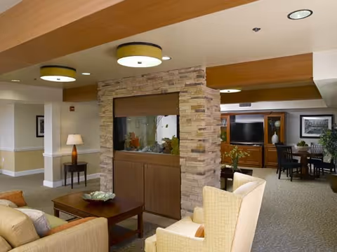 Communal living room with upholstered chairs and sofa, a central stone-encased aquarium, coffee table, and dining area visible in the background.
