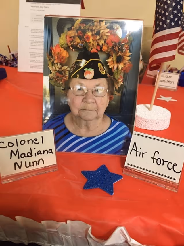 A framed photograph of an elderly woman wearing glasses and a decorated military hat with a peach emblem, placed on a red tablecloth. In front of the photo are two small signs, one reading 'Colonel Madiana Nunn' and the other 'Air force'. There is a small American flag on a stand to the right and a blue glittery star decoration on the table.