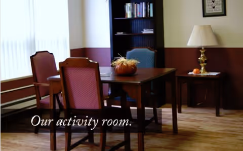 A room with a wooden table surrounded by four chairs, a bookshelf in the background, a side table with a lamp and a small decorative item, and a window with vertical blinds letting in natural light. The text 'Our activity room.' is overlaid on the image.