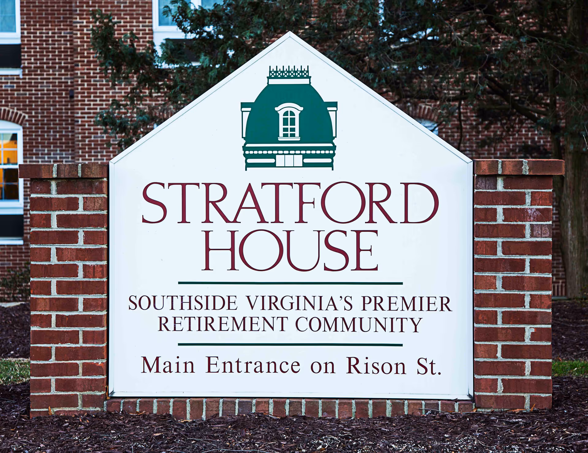 A large white sign with a green graphic of a building roof and the text 'STRATFORD HOUSE Southside Virginia's Premier Retirement Community Main Entrance on Rison St.' The sign is supported by two brick pillars and is set in front of a brick building with windows and some trees.