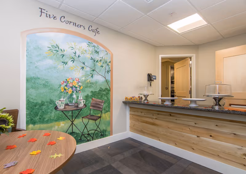 Interior view of a cafe area named Five Corners Cafe with a mural on the wall depicting a small table with a vase of flowers and two chairs. There is a wooden counter with cake stands and a door leading to another room. A round table with artificial autumn leaves is partially visible in the foreground.