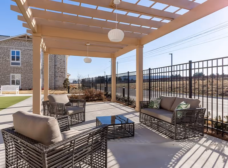 Outdoor seating area under a pergola with cushioned wicker chairs and a sofa arranged around a glass-top coffee table, next to a black metal fence with a building and open field in the background.