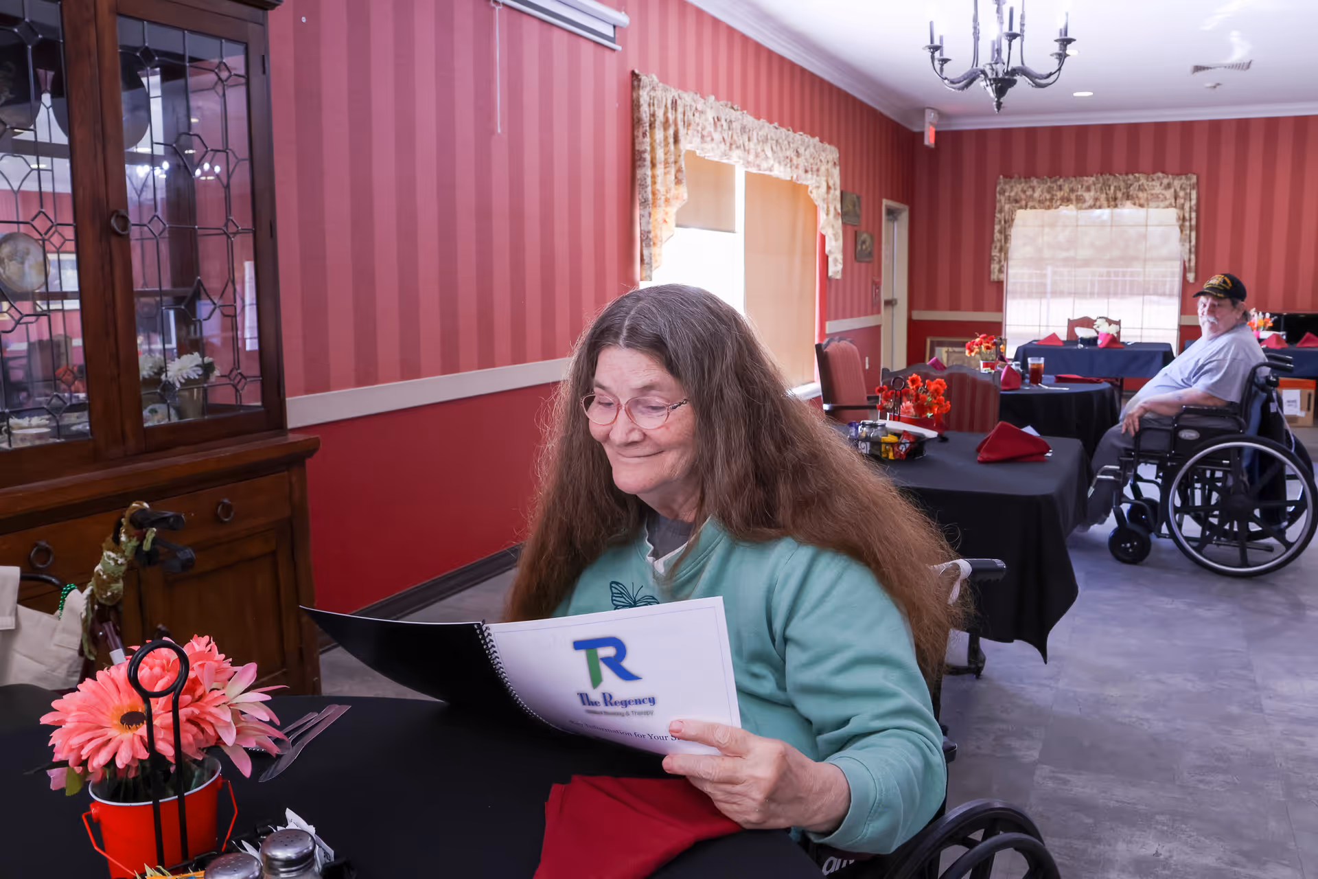 Two elderly individuals in wheelchairs are seated at separate tables in a dining room with red striped wallpaper and floral curtains. One woman with long hair and glasses is smiling while looking at a menu from The Regency Skilled Nursing & Therapy. The tables are covered with black tablecloths and decorated with red napkins and flower arrangements.