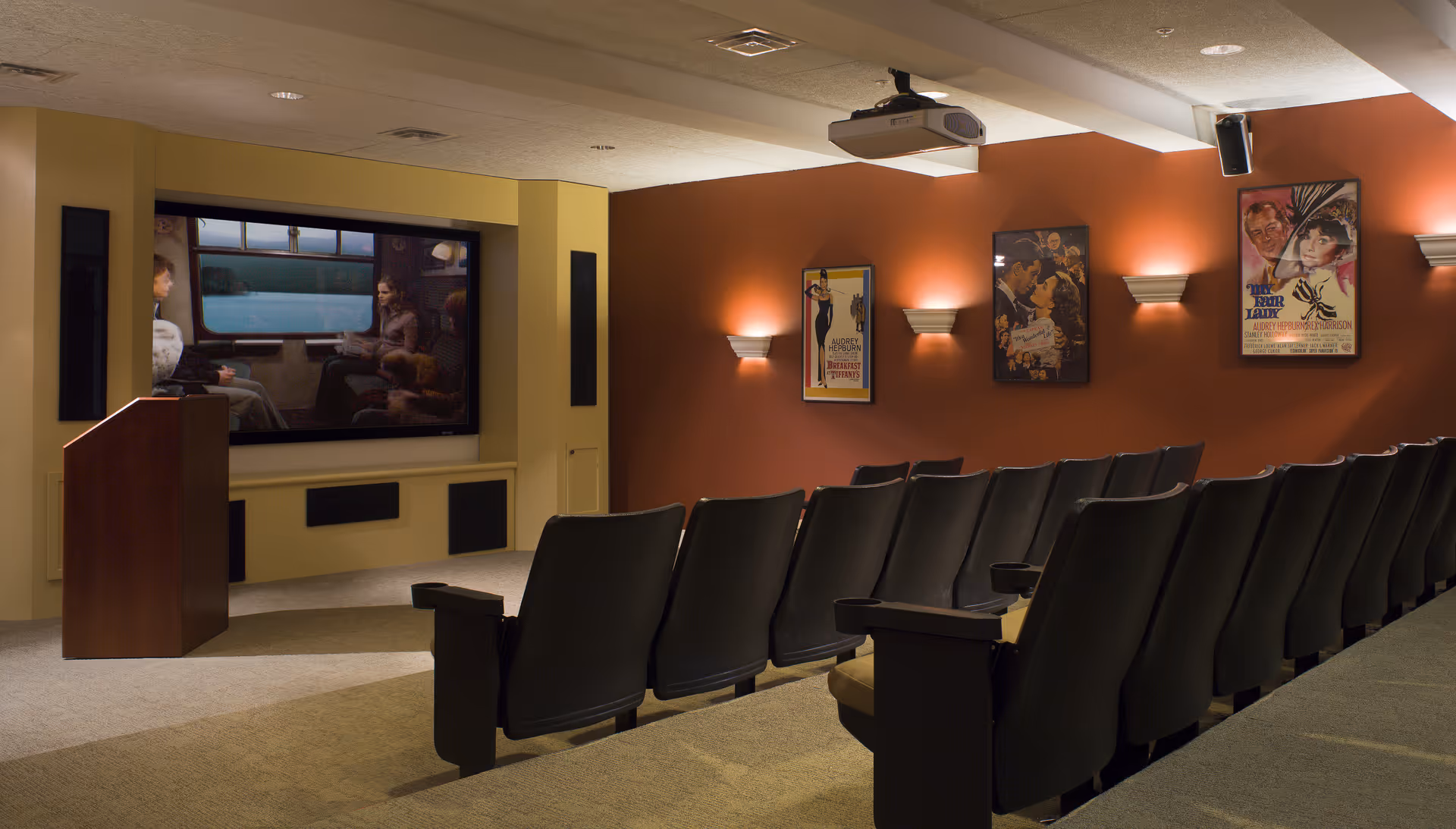 A small theater room with rows of black cushioned seats facing a large screen displaying a movie scene. The walls are painted a warm brown color and decorated with framed vintage movie posters. There is a wooden podium to the left of the screen and a ceiling-mounted projector above.