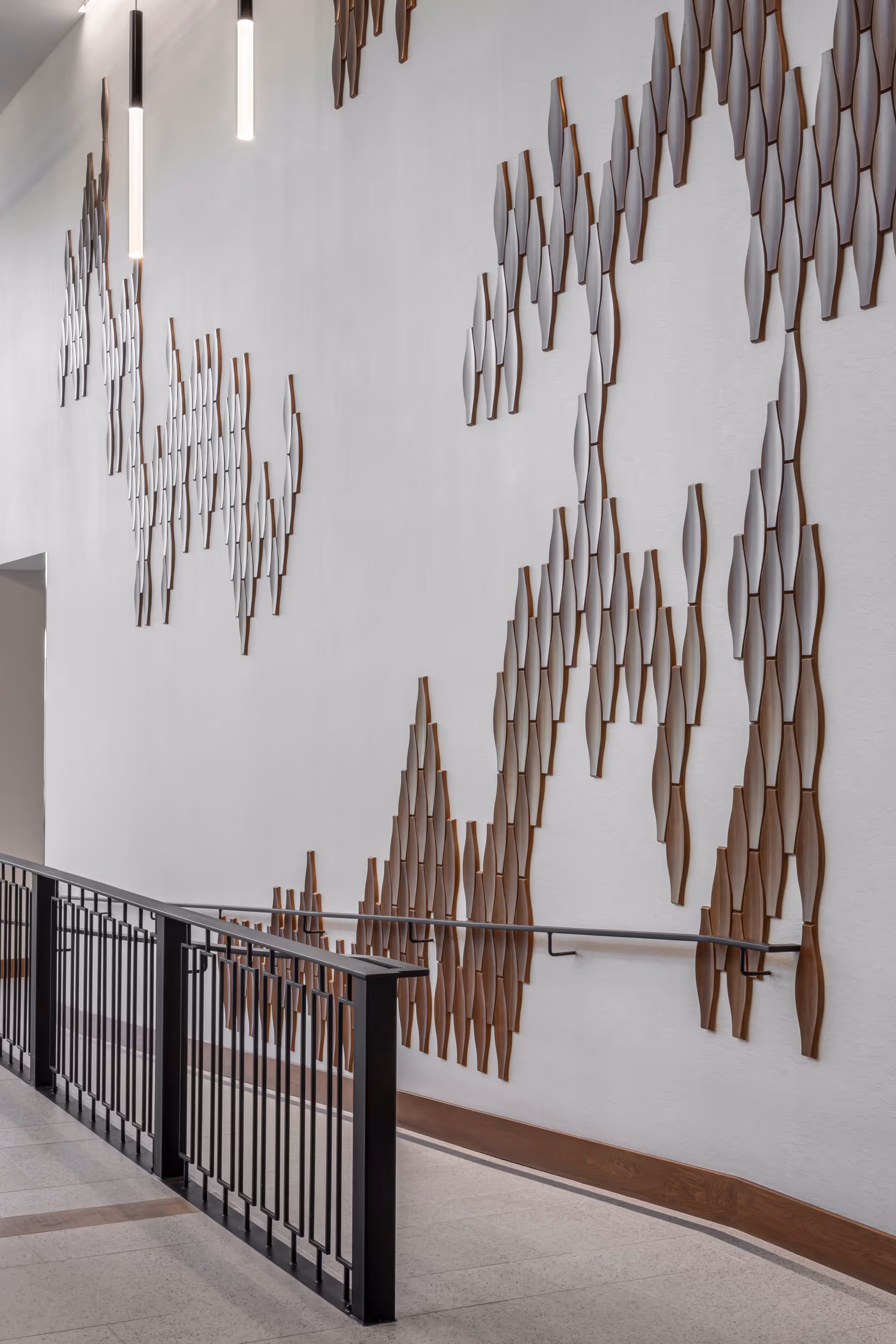 Interior view of a hallway with a black metal railing and a wall decorated with abstract wooden art pieces. The wall art consists of multiple elongated wooden shapes arranged in a flowing pattern. Two modern pendant lights hang from the ceiling.