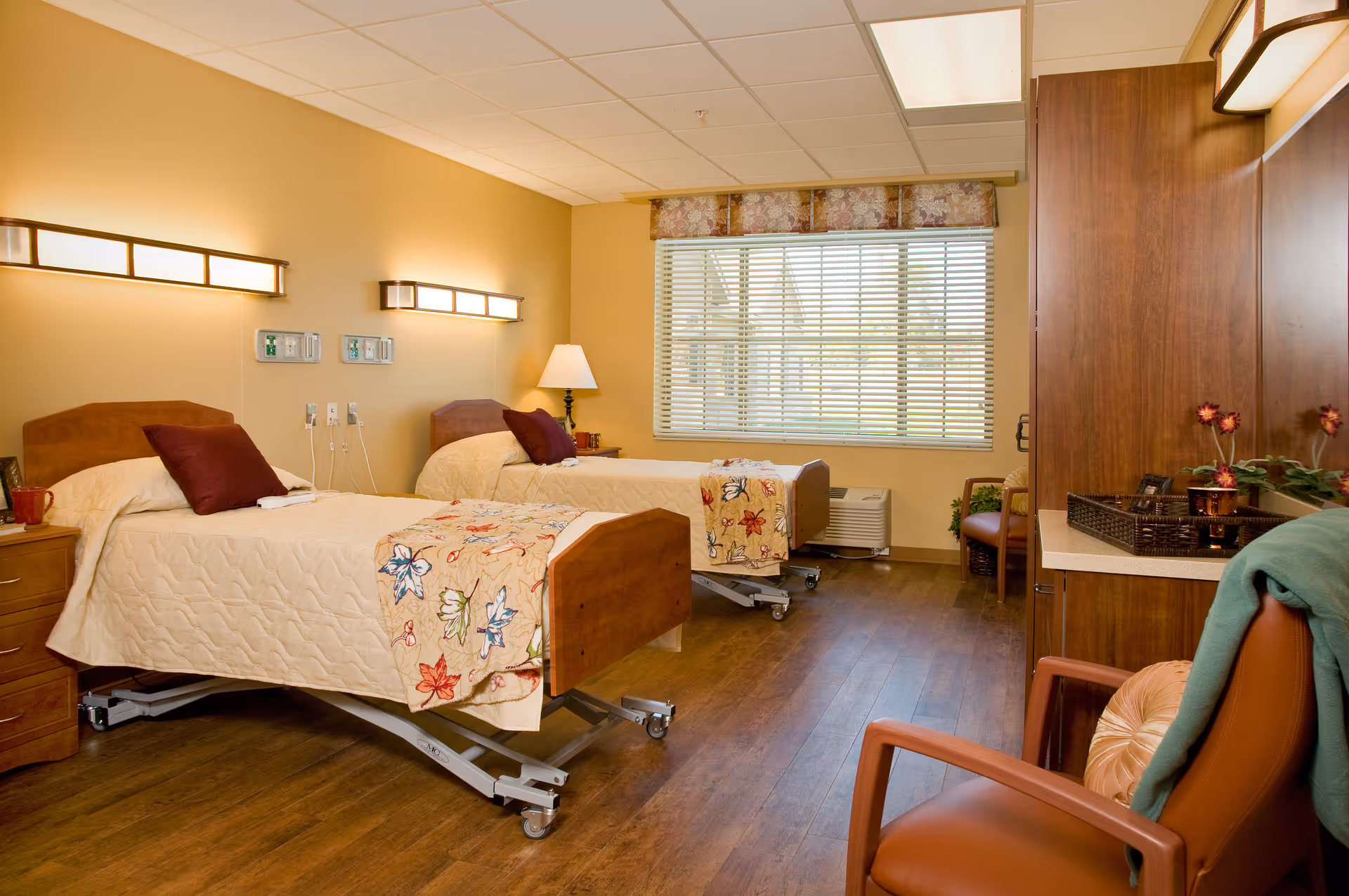 A well-lit senior living bedroom with two hospital-style beds, each with a beige quilt and a decorative pillow. The room has wooden flooring, a large window with blinds, a bedside table with a lamp, and a comfortable chair with a blanket draped over it. There is also a wooden cabinet with decorative items on top.