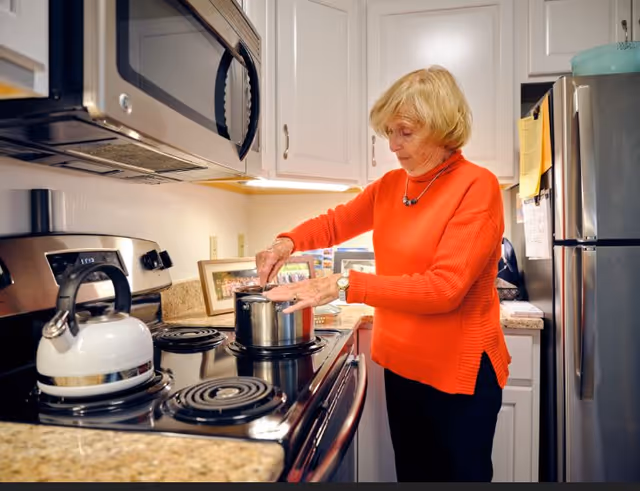 An elderly woman wearing an orange sweater is cooking in a kitchen. She is stirring a pot on a stove with a kettle nearby. The kitchen has white cabinets, a microwave above the stove, and a stainless steel refrigerator.