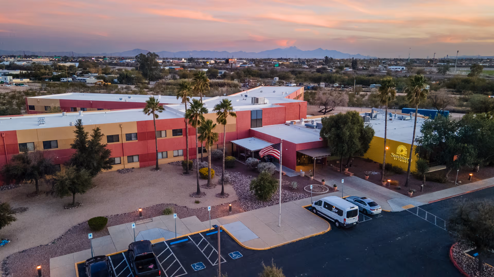 Aerial view of a senior living facility building with a red and beige exterior, surrounded by desert landscaping with palm trees and other vegetation. There is a parking lot with several vehicles, including a white van, and an American flag flying on a flagpole near the entrance. The sky is at sunset with soft pink and orange hues, and mountains are visible in the distance.