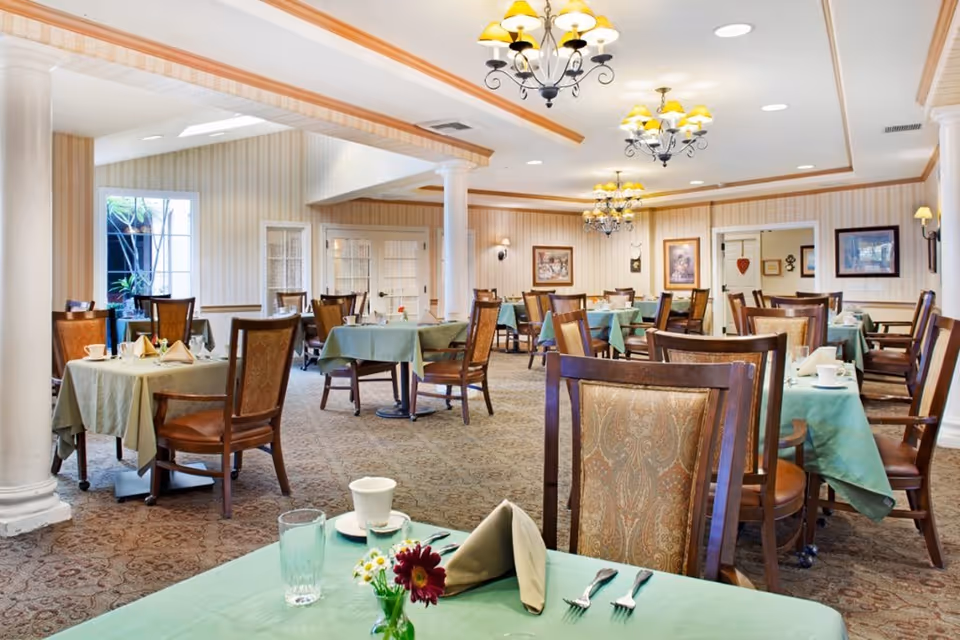 A spacious dining room with multiple tables covered in green tablecloths, each set with cups, glasses, napkins, and silverware. The room features wooden chairs with patterned upholstery, chandeliers with yellow lampshades hanging from the ceiling, and beige patterned carpet. The walls are decorated with framed pictures and have a striped wallpaper design. Large windows and columns are also visible.