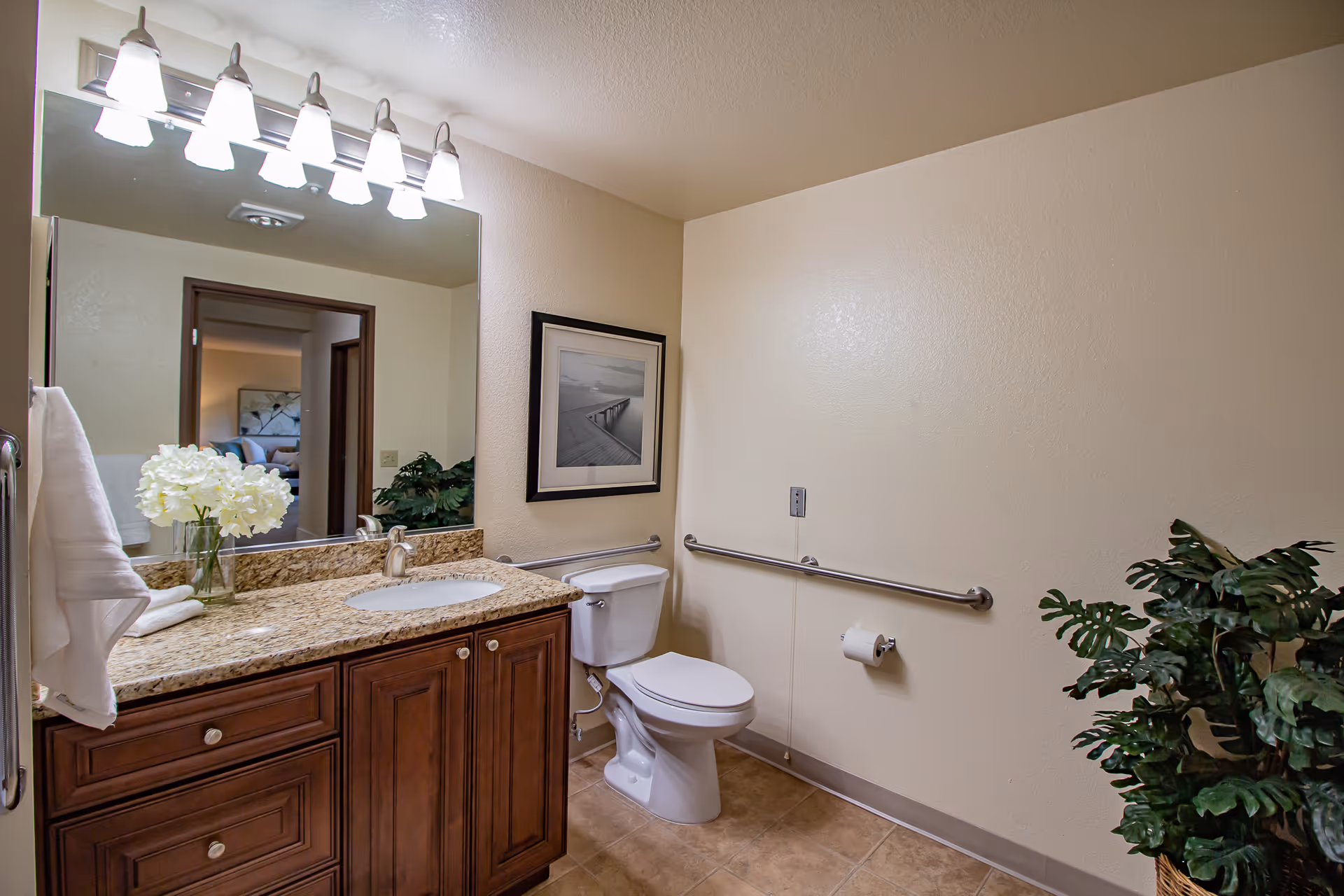 A clean and well-lit bathroom featuring a granite countertop with a sink, a large mirror with six light fixtures above it, a white toilet with a grab bar on the wall beside it, a framed black and white photo on the wall, a towel hanging on a rack, a vase with white flowers on the counter, and a green potted plant on the floor.