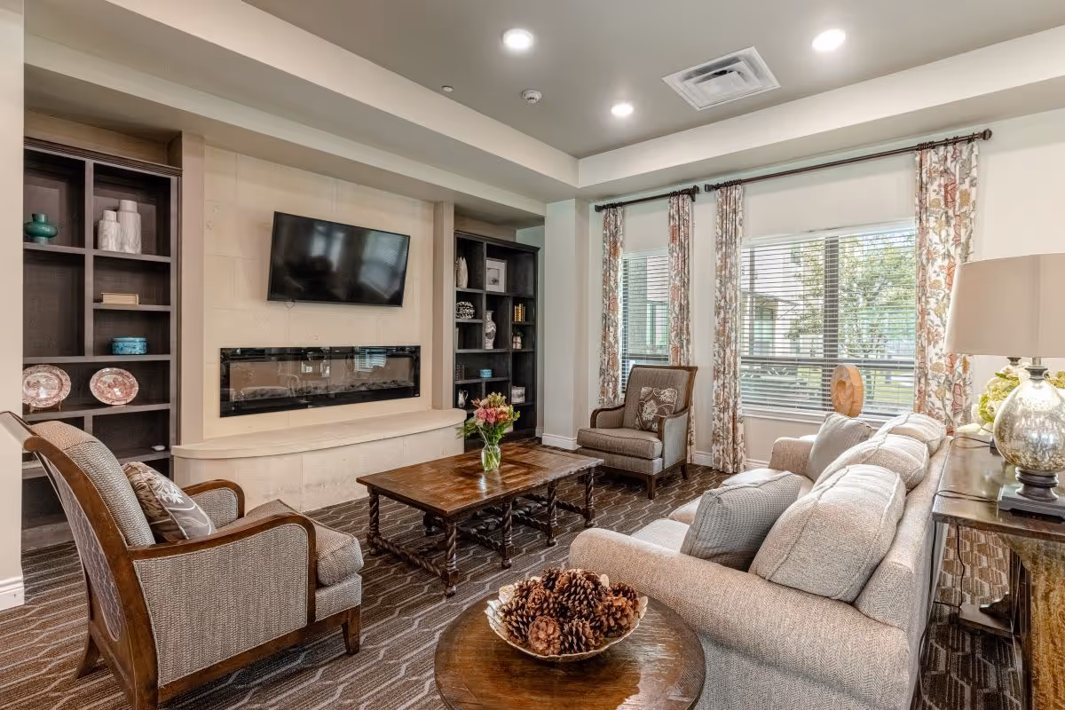A cozy living room with a beige sofa, two armchairs, a wooden coffee table with a vase of flowers, and a round side table with a bowl of pinecones. The room features a wall-mounted flat-screen TV above a modern electric fireplace, built-in dark wood shelves with decorative items, large windows with floral curtains, and a table lamp on a side table.