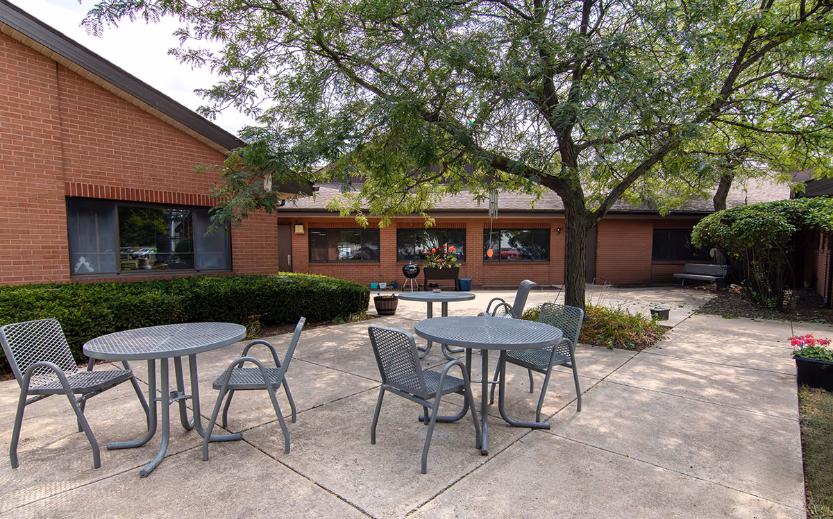 Outdoor patio area at Renwick Nursing & Rehab with metal tables and chairs on a concrete surface, surrounded by brick buildings and greenery including a large tree providing shade.