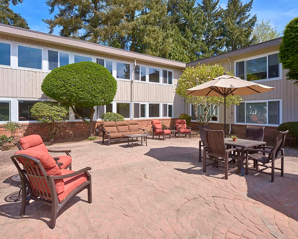 Sunny courtyard patio with outdoor seating, tables, and an umbrella in front of a two-story senior living building.
