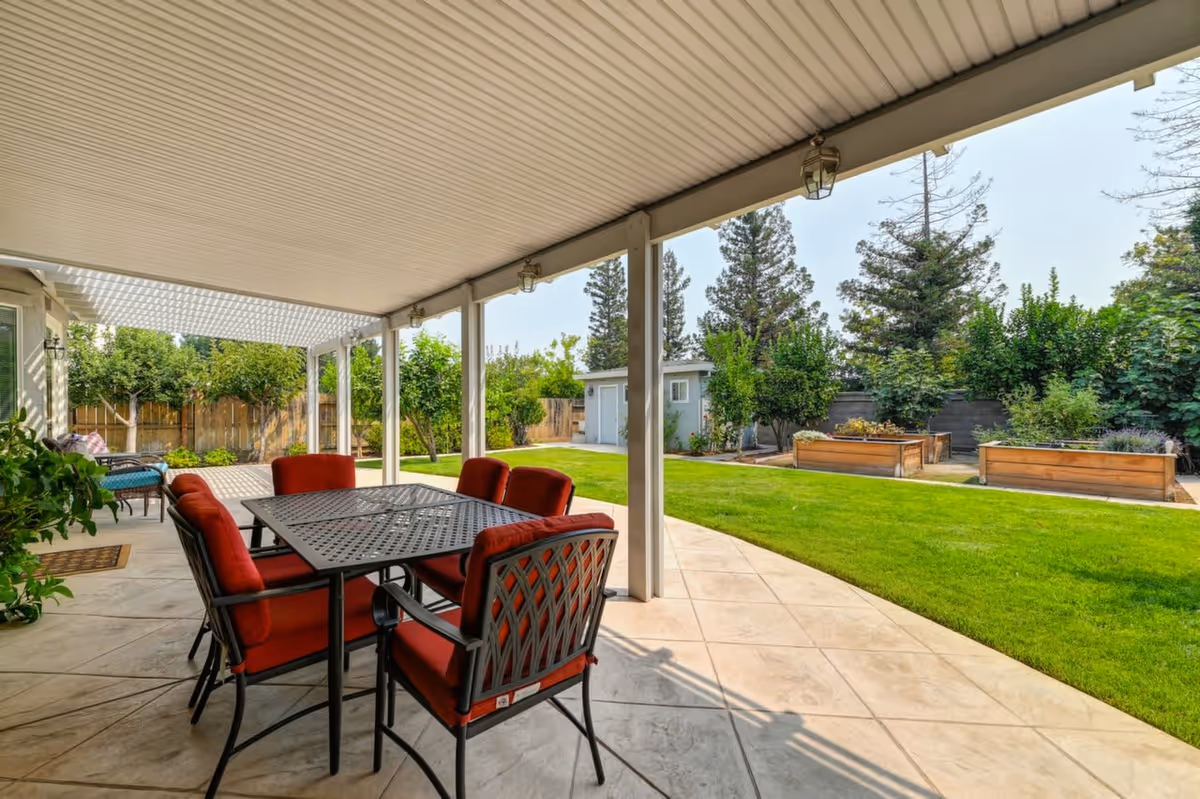 Covered outdoor patio area with a black metal table and six red cushioned chairs. The patio overlooks a well-maintained green lawn with raised garden beds and various trees and shrubs. A small shed is visible in the background under a clear sky.