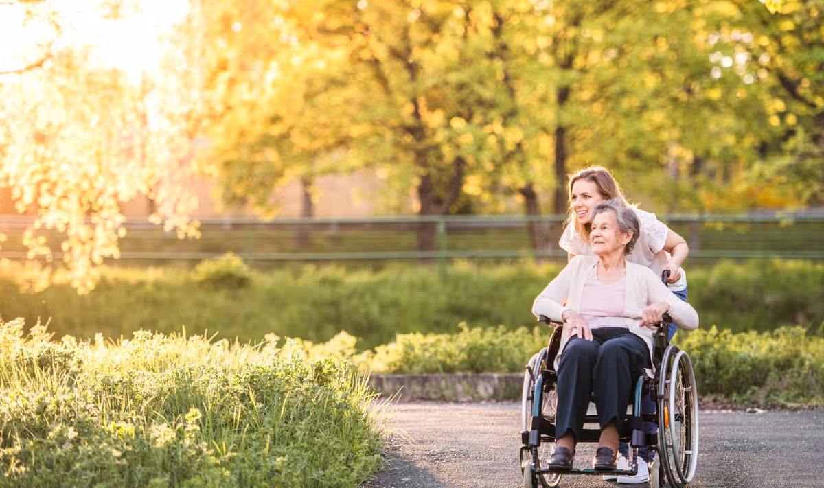 A young woman pushing an elderly woman in a wheelchair along a paved path surrounded by green grass and trees with sunlight filtering through the leaves.