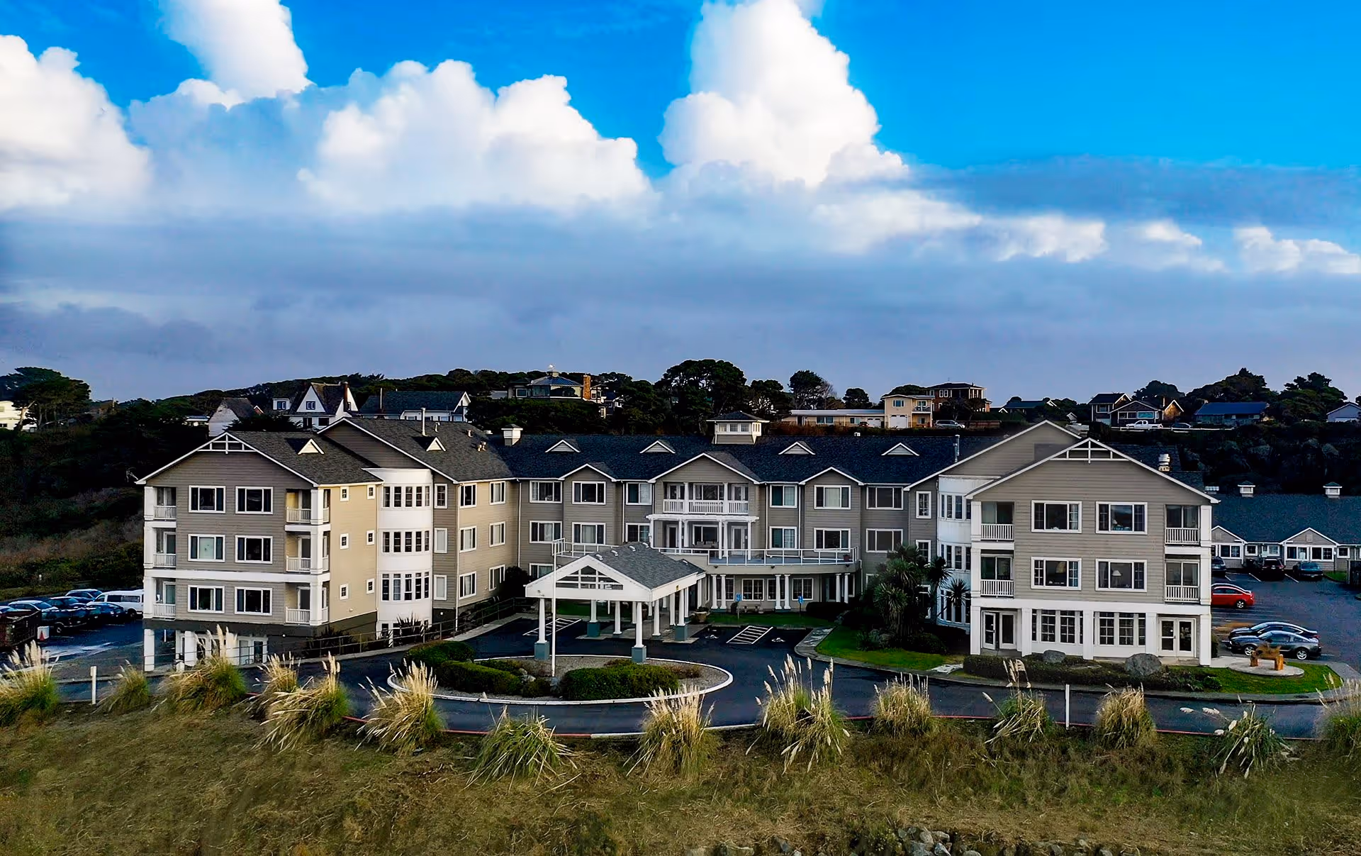Exterior view of a large, multi-story senior living facility building with beige siding and white trim, surrounded by greenery and parked cars under a partly cloudy blue sky.