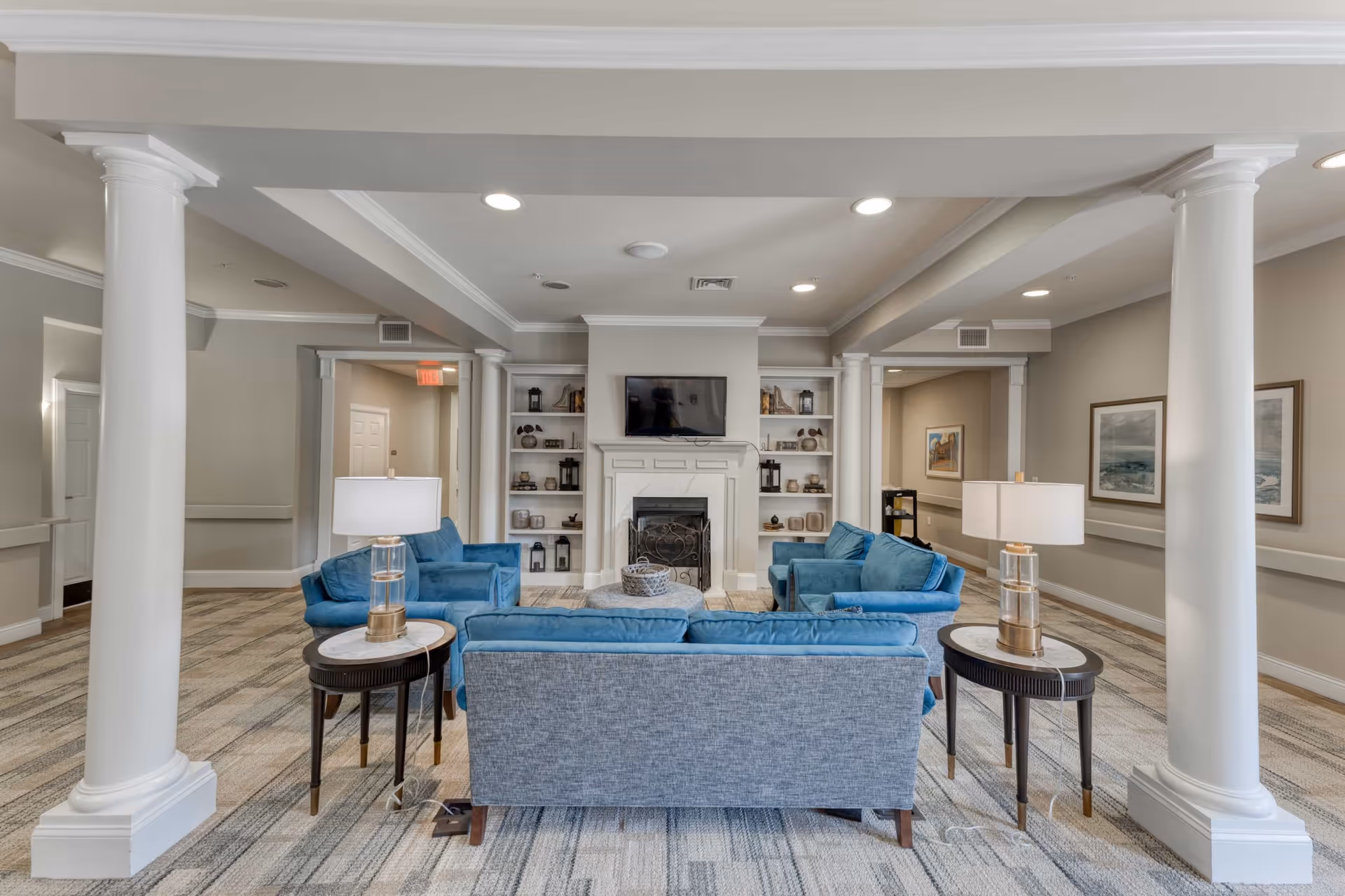 A spacious and well-lit living room area with blue upholstered sofas arranged around a central fireplace. The fireplace is flanked by built-in white shelves decorated with various ornaments. Two side tables with lamps are positioned beside the sofas. The room features white columns, neutral-colored walls, framed artwork, and recessed ceiling lights.