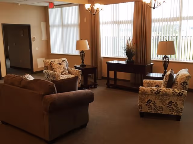 A cozy living room area with a brown sofa, two floral-patterned armchairs, two side tables with lamps, a console table with a plant, and large windows with vertical blinds letting in natural light.