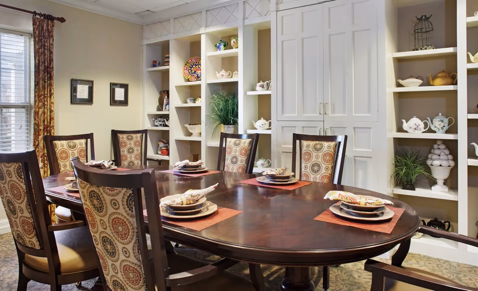 A dining room with a large oval wooden table set for six people with plates, napkins, and placemats. The chairs have patterned upholstery. Behind the table are built-in white shelves displaying decorative teapots, plants, and other ornaments. There are two framed pictures on the wall and a window with patterned curtains.