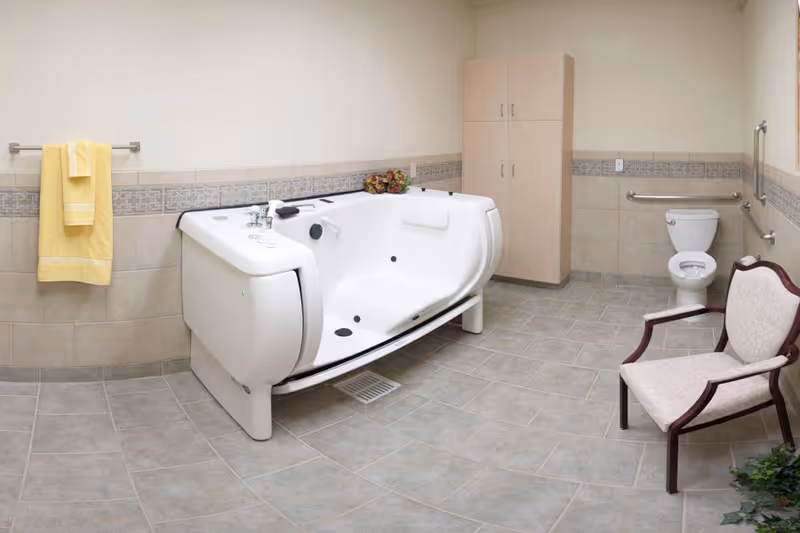 Accessible senior bathroom featuring a white walk-in bathtub, toilet with grab bars, a towel on a rack, a storage cabinet and a chair.